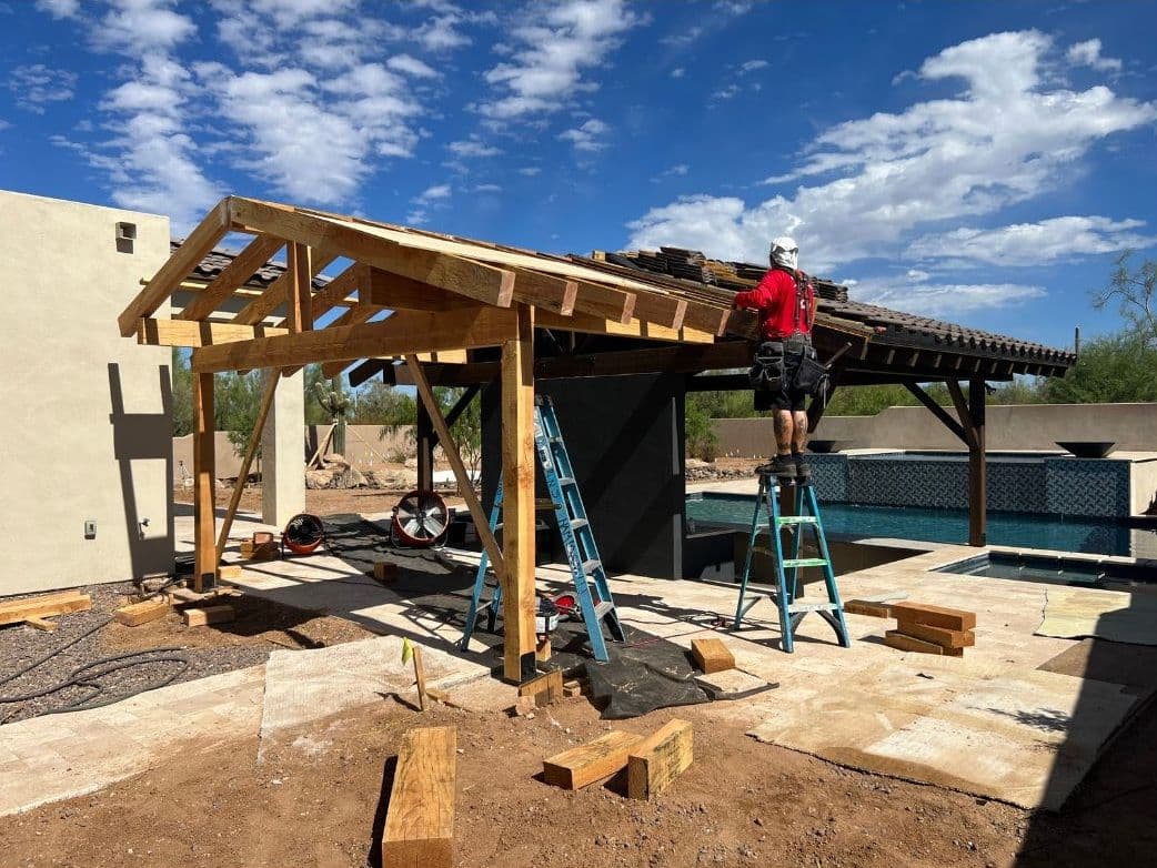 Construction worker building a wooden pergola over a poolside patio in sunny outdoor setting.