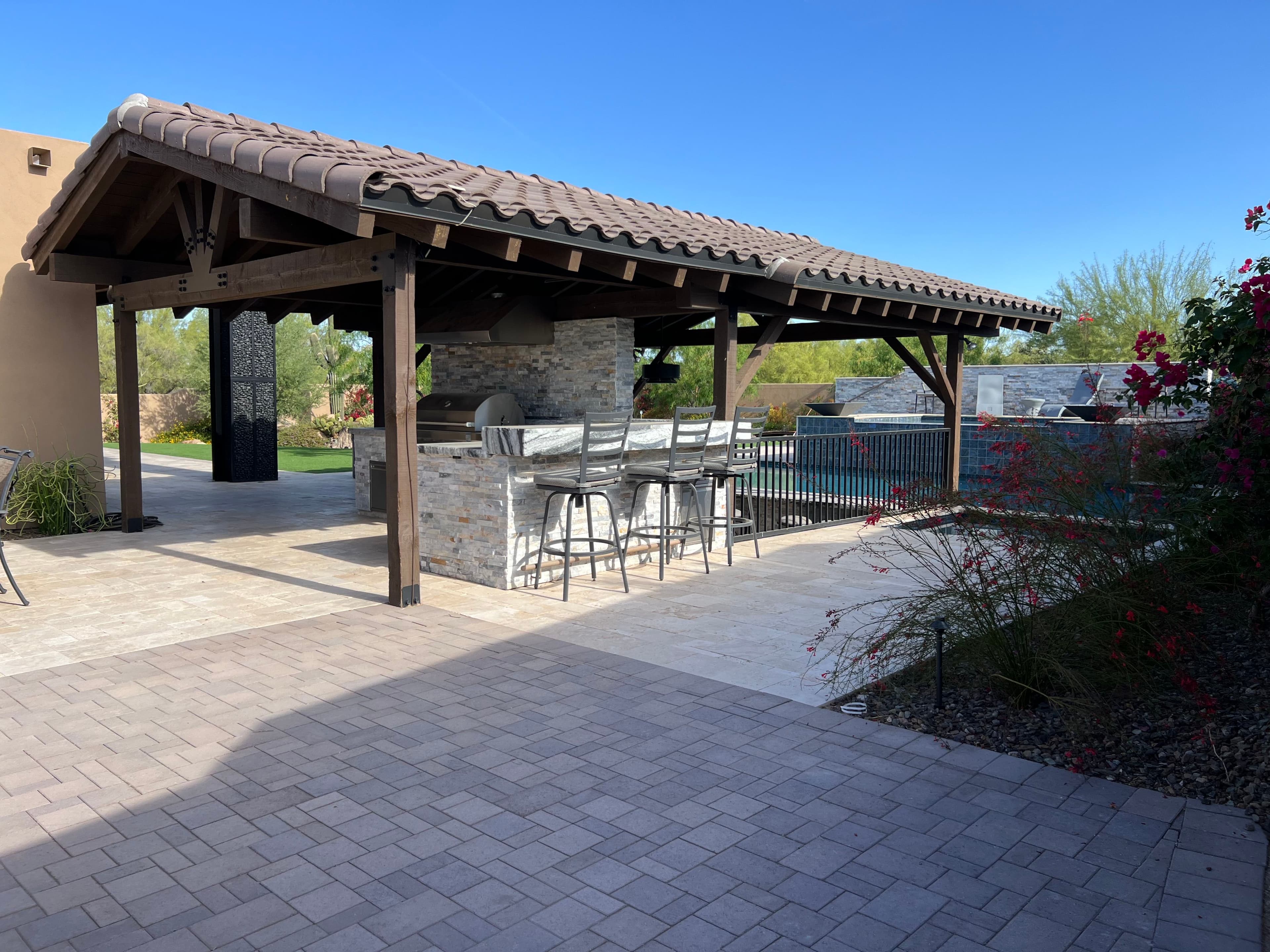 Modern outdoor kitchen with bar seating under a wooden pergola, surrounded by landscaping.