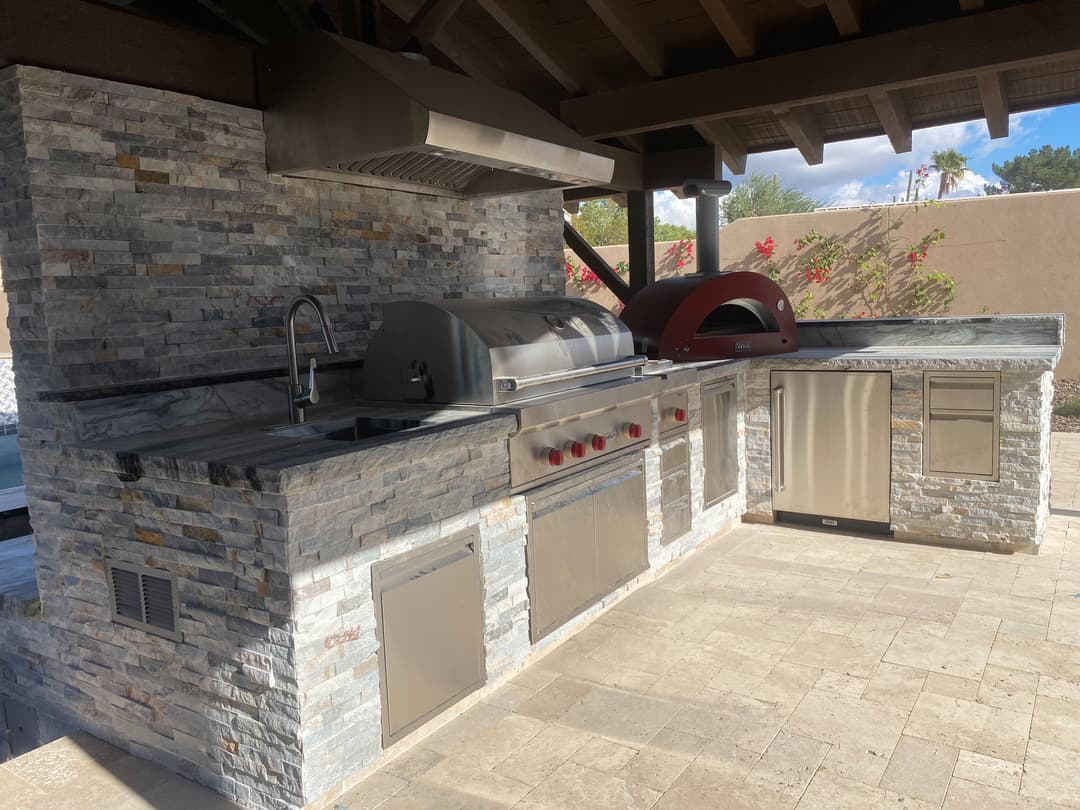 Outdoor kitchen with stone veneer, grill, pizza oven, and stainless steel appliances.