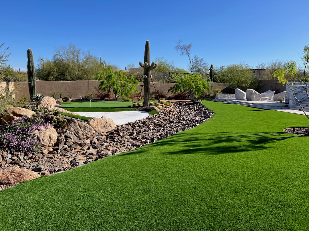 Lush green artificial turf landscaping with rocks and cactus under a clear blue sky.